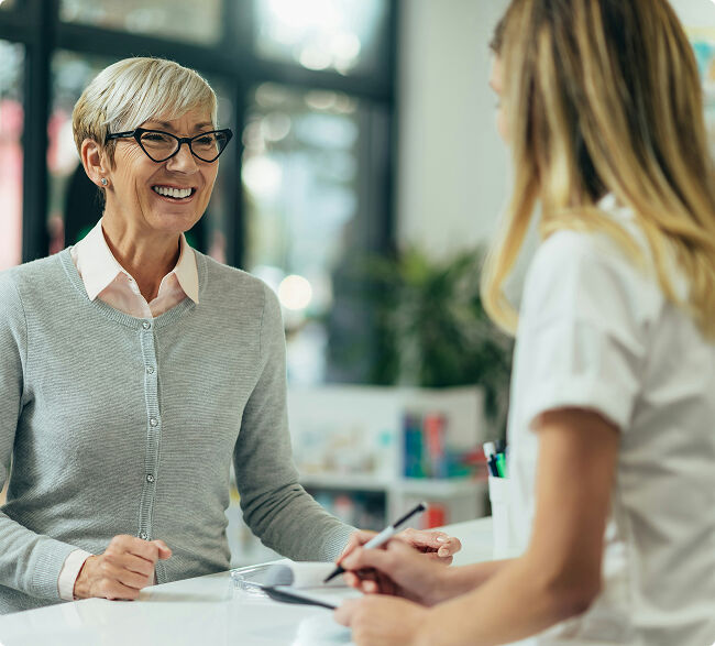 A smiling older woman wearing glasses talking with a healthcare professional across a clinic counter